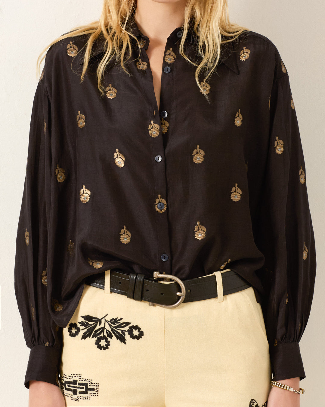 Wearing the Emery Shadow Bamboo Shirt, a black long-sleeve with vintage appeal and metallic gold florals, tucked into high-waisted cream pants with black embroidery and finished with a black belt.