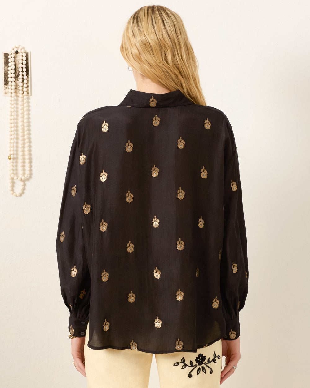 A woman with long blonde hair, seen from the back beside hanging pearls, wears the Emery Shadow Bamboo Shirt in black featuring a metallic gold floral motif.