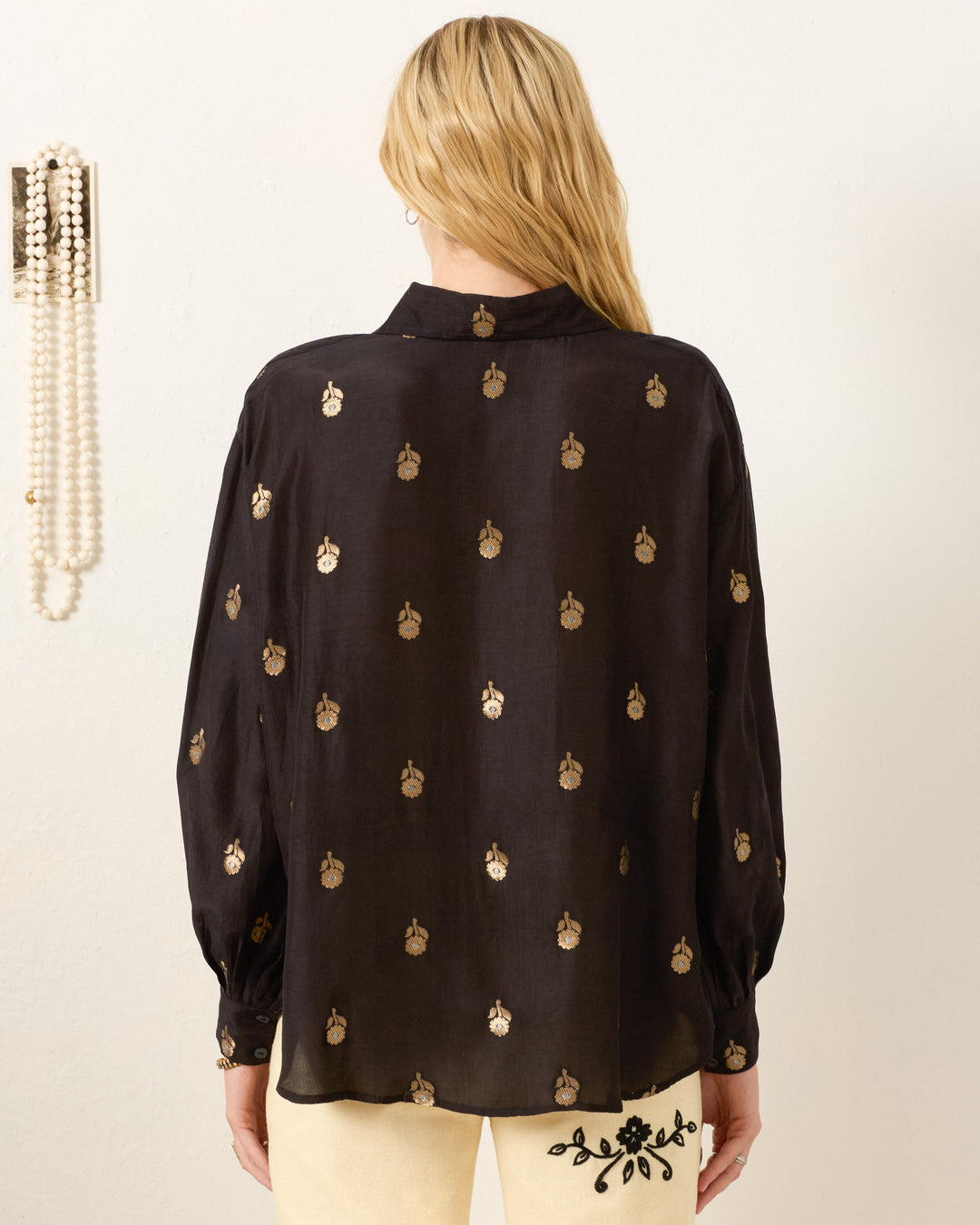 A woman with long blonde hair, seen from the back beside hanging pearls, wears the Emery Shadow Bamboo Shirt in black featuring a metallic gold floral motif.