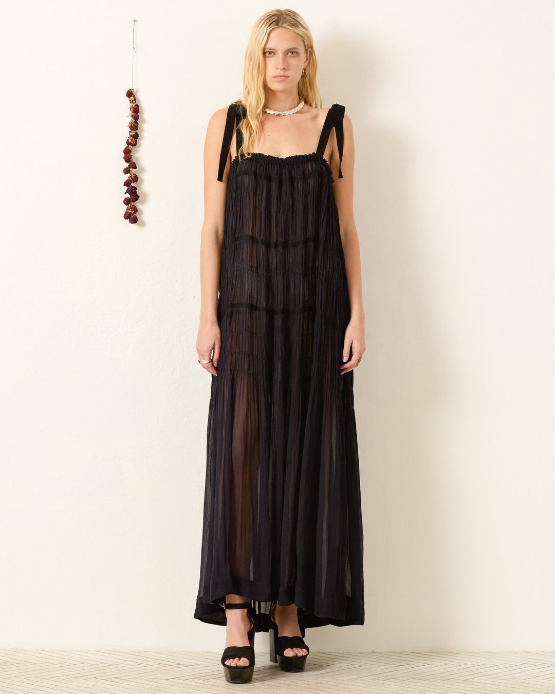 A person stands against a plain light wall wearing the Emrys Shadow Chiffon Dress—sheer black cupro-chiffon with shoulder ties and ruched tiers—paired with black platform heels. A string of dried red peppers hangs to the left.