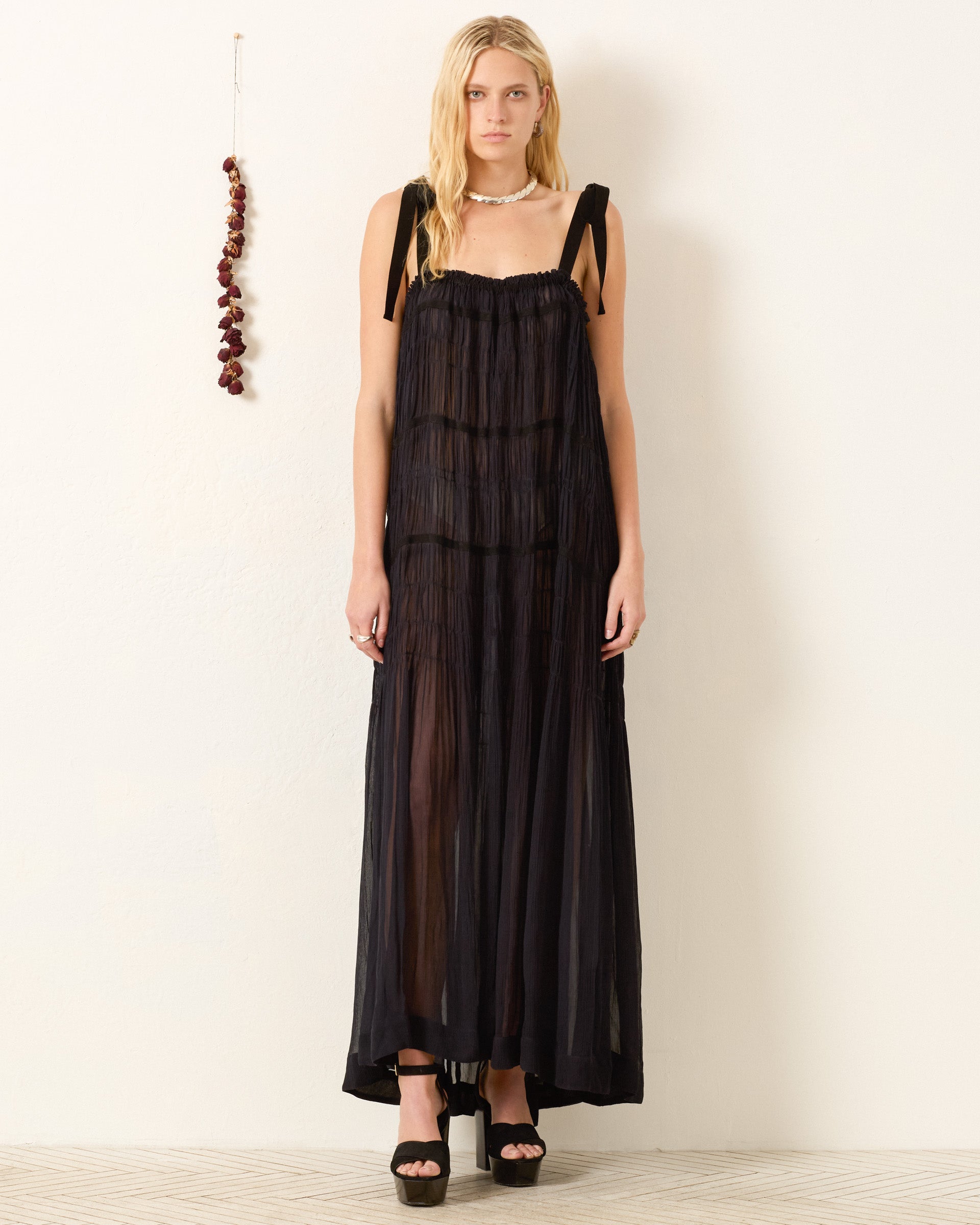 A person stands against a plain light wall wearing the Emrys Shadow Chiffon Dress—sheer black cupro-chiffon with shoulder ties and ruched tiers—paired with black platform heels. A string of dried red peppers hangs to the left.