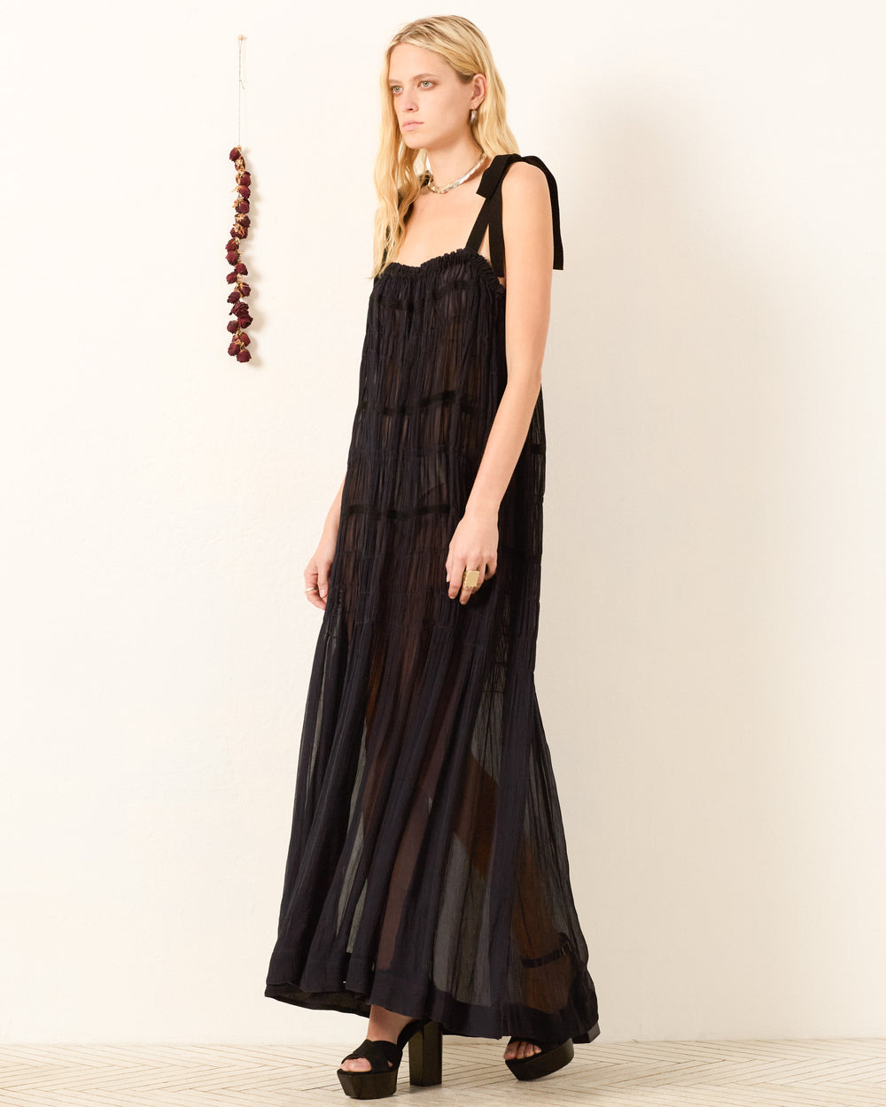 A woman stands against a plain wall with a string of dried flowers, wearing the Emrys Shadow Chiffon Dress—a long, sheer black dress with shoulder ties and ruched tiers—paired with black platform sandals.