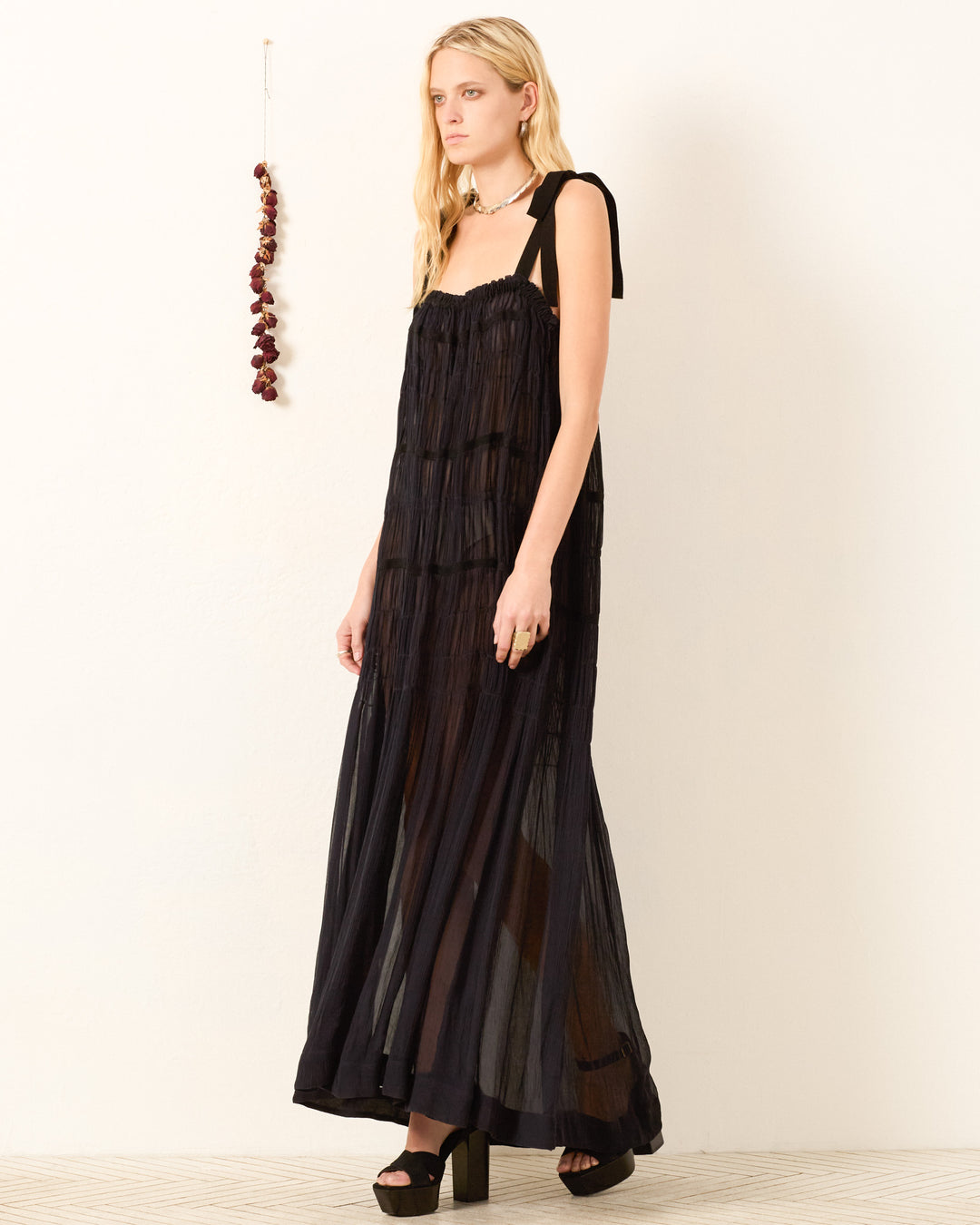 A woman stands against a plain wall with a string of dried flowers, wearing the Emrys Shadow Chiffon Dress—a long, sheer black dress with shoulder ties and ruched tiers—paired with black platform sandals.