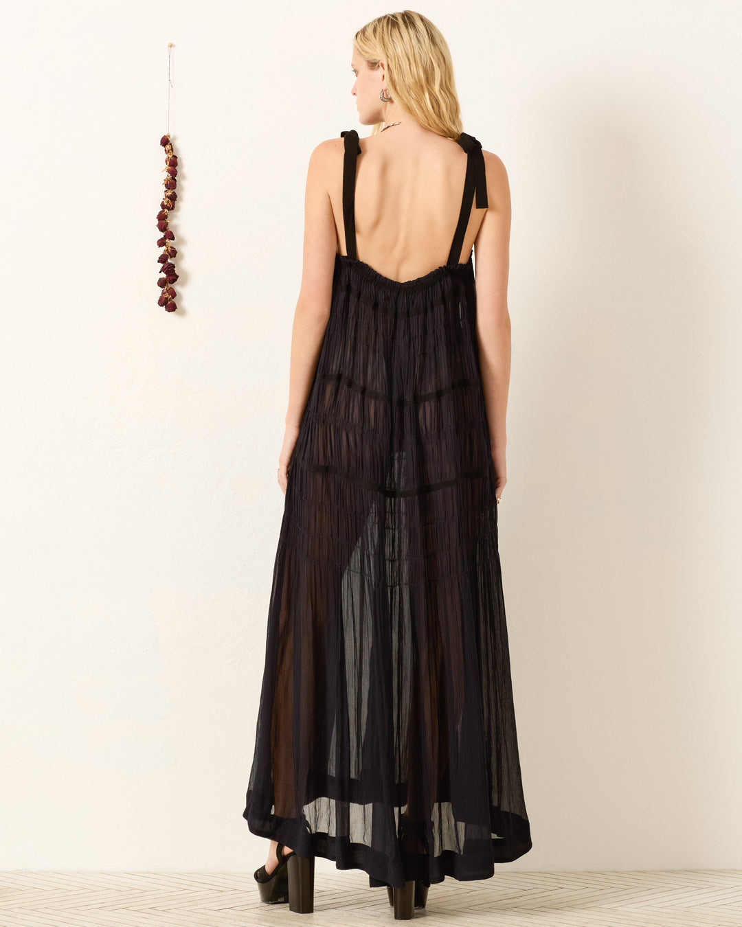 A woman, her back exposed by slender shoulder straps, wears the Emrys Shadow Chiffon Dress in sheer black cupro-chiffon. She stands by a white wall decorated with a string of dried red peppers.