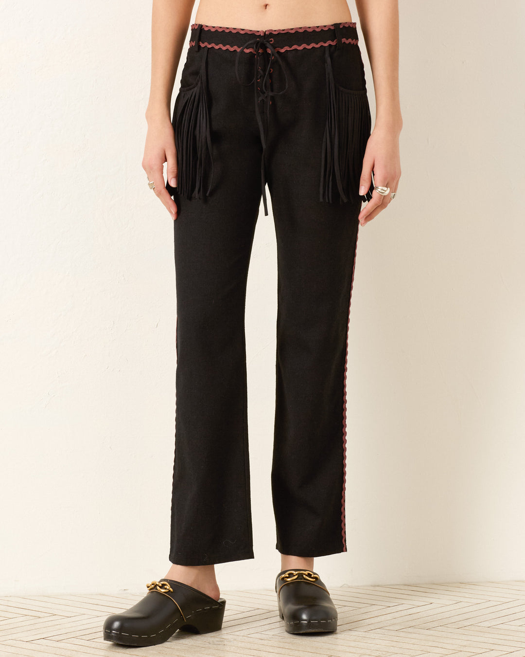 A person stands against a light wall wearing the Ezra Noir Rein Pant—black lace-up pants with fringe on the front pockets, red side trim—and black clogs with gold detailing.