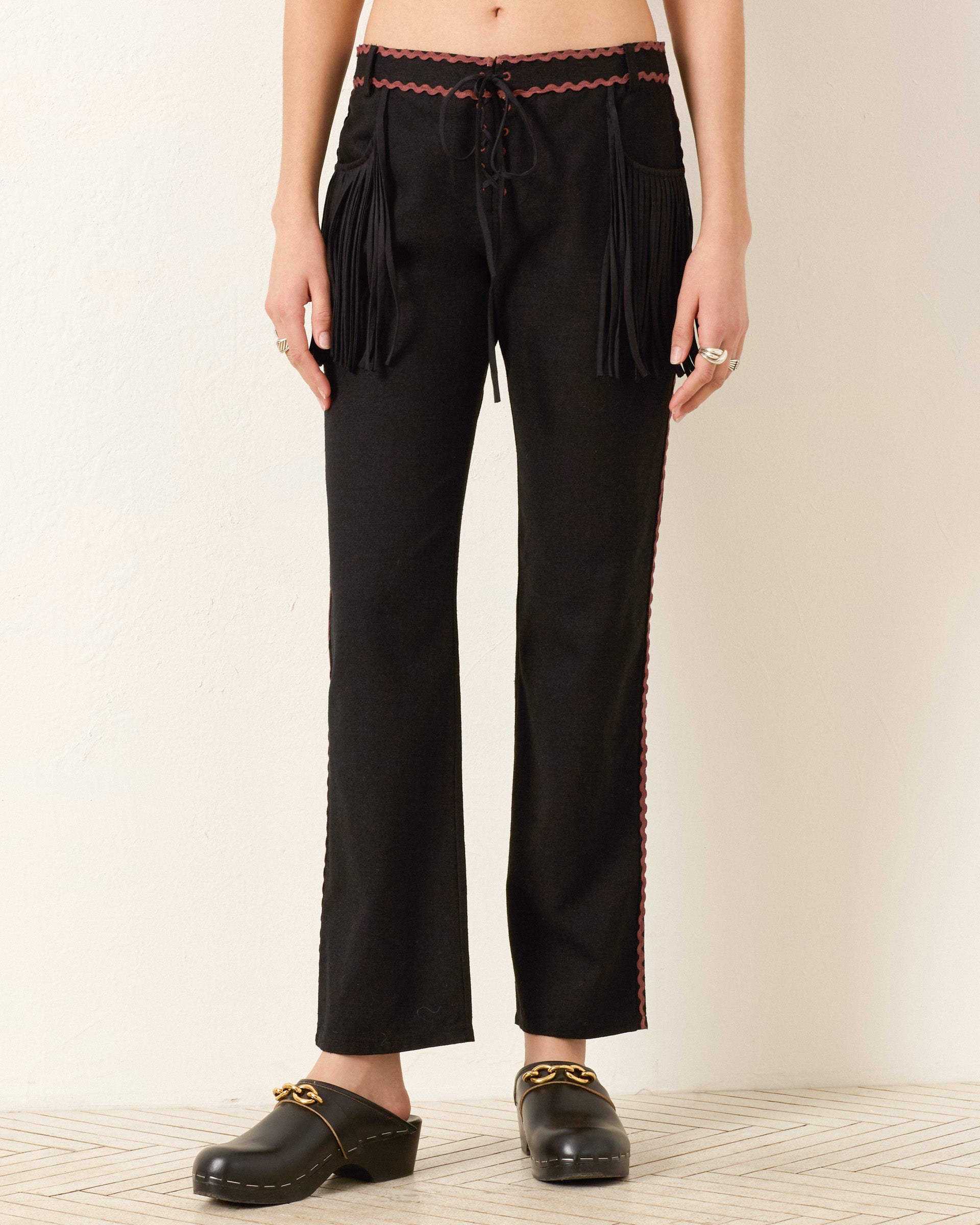 A person stands against a light wall wearing the Ezra Noir Rein Pant—black lace-up pants with fringe on the front pockets, red side trim—and black clogs with gold detailing.