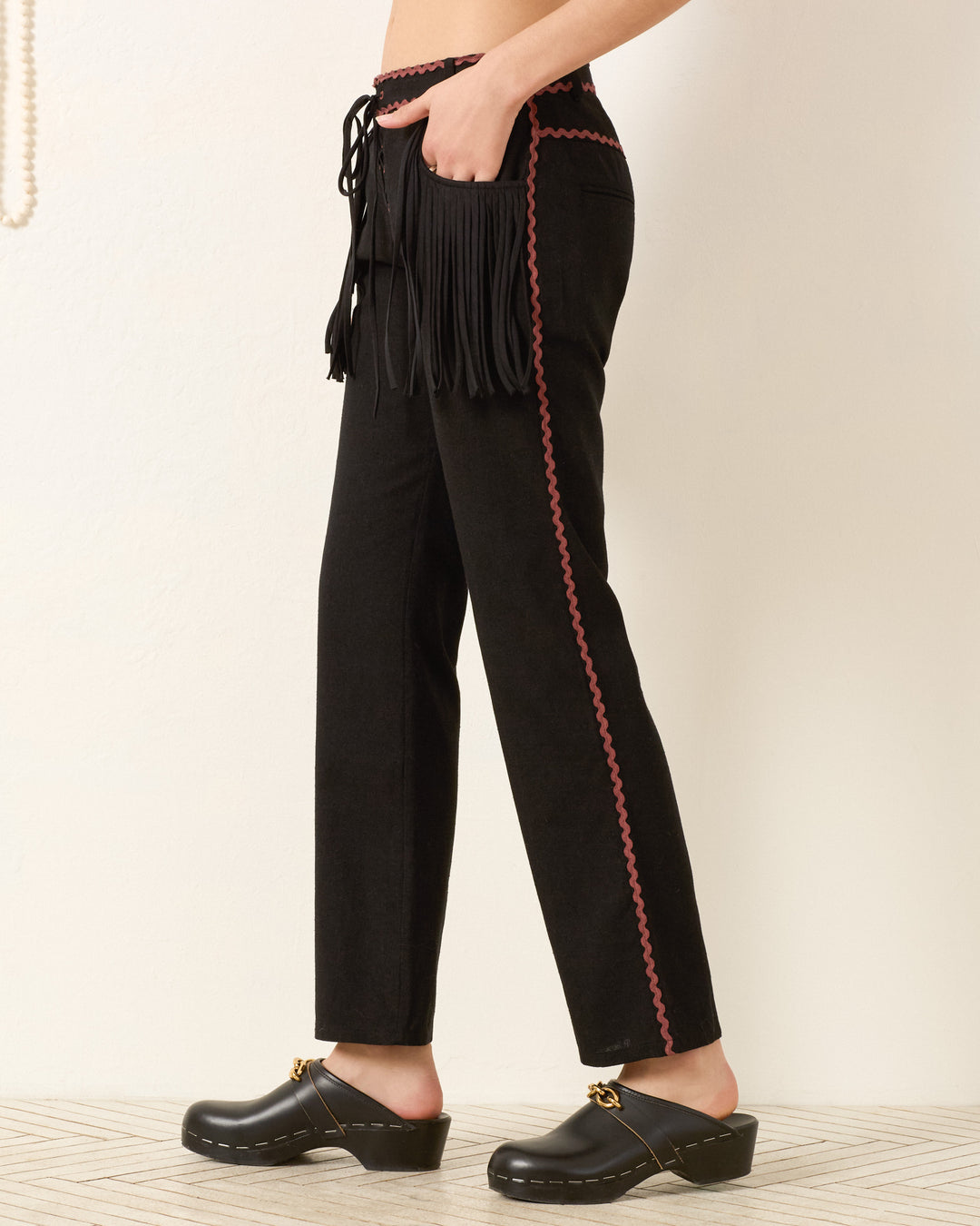A person wears the Ezra Noir Rein Pant, featuring a classic straight-leg silhouette with black fabric, pink trim, and fringe detail, paired with black clogs and standing against a neutral background.