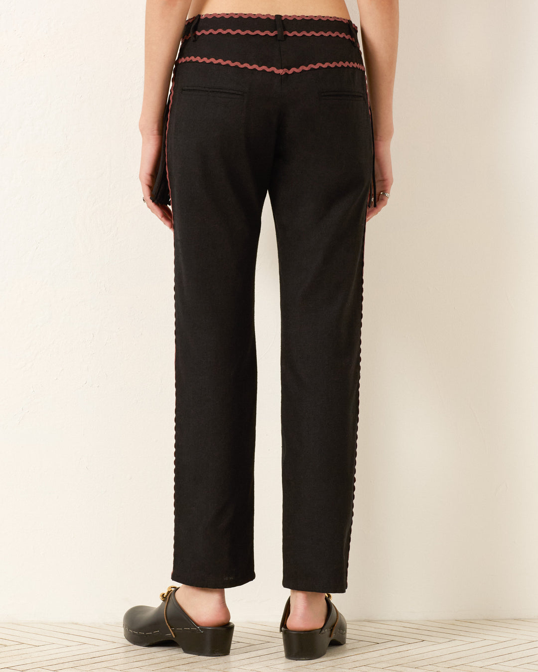 A person faces away, wearing the Ezra Noir Rein Pant—black straight-leg pants with red decorative trim—paired with black clogs, standing against a plain light background.