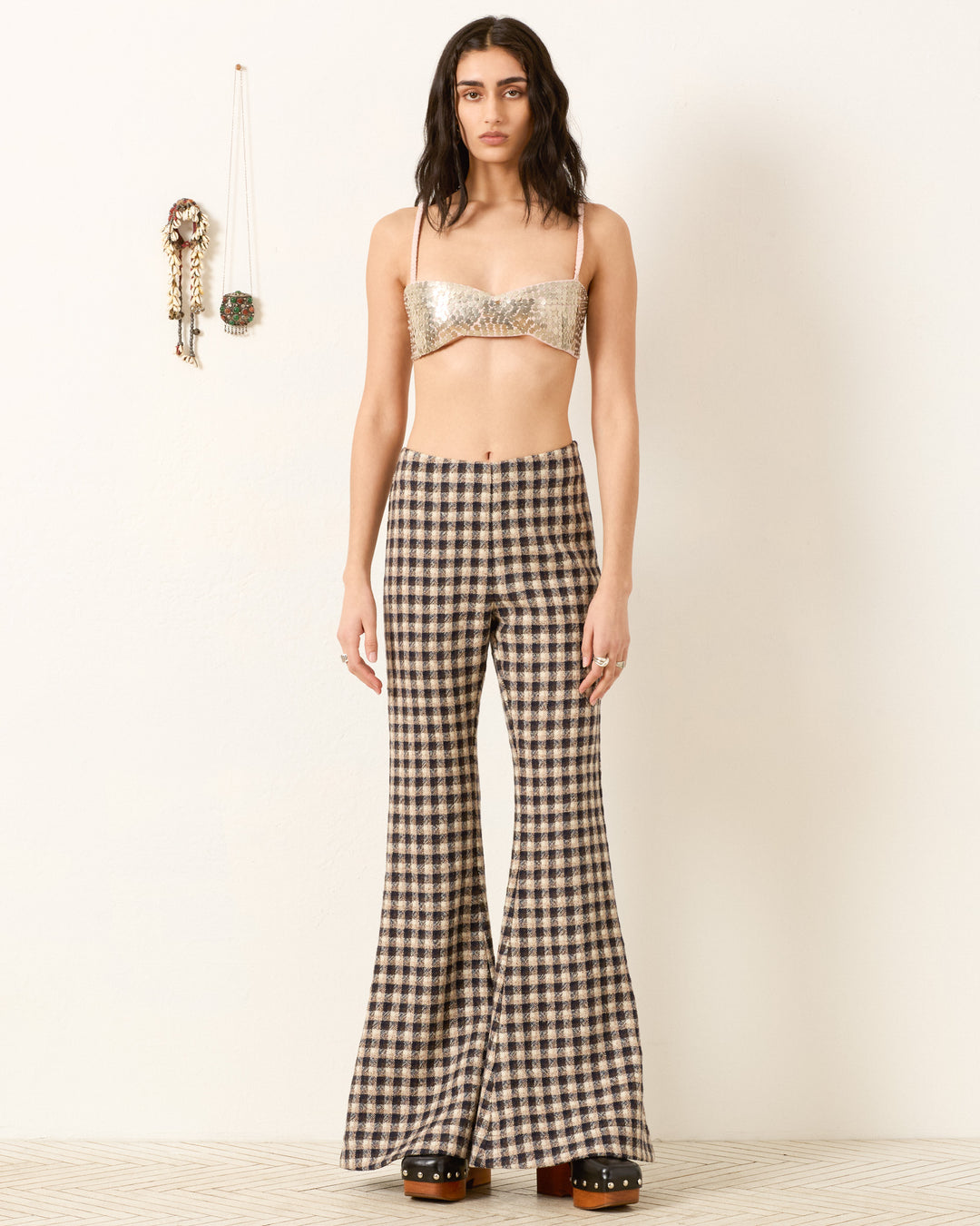 A person stands against a plain wall, wearing a metallic bralette and the Farah Trail Dust Pant—a plaid, high-waisted, bell-bottom style crafted from luxurious Italian virgin wool. Decor hangs on the wall to the left.
