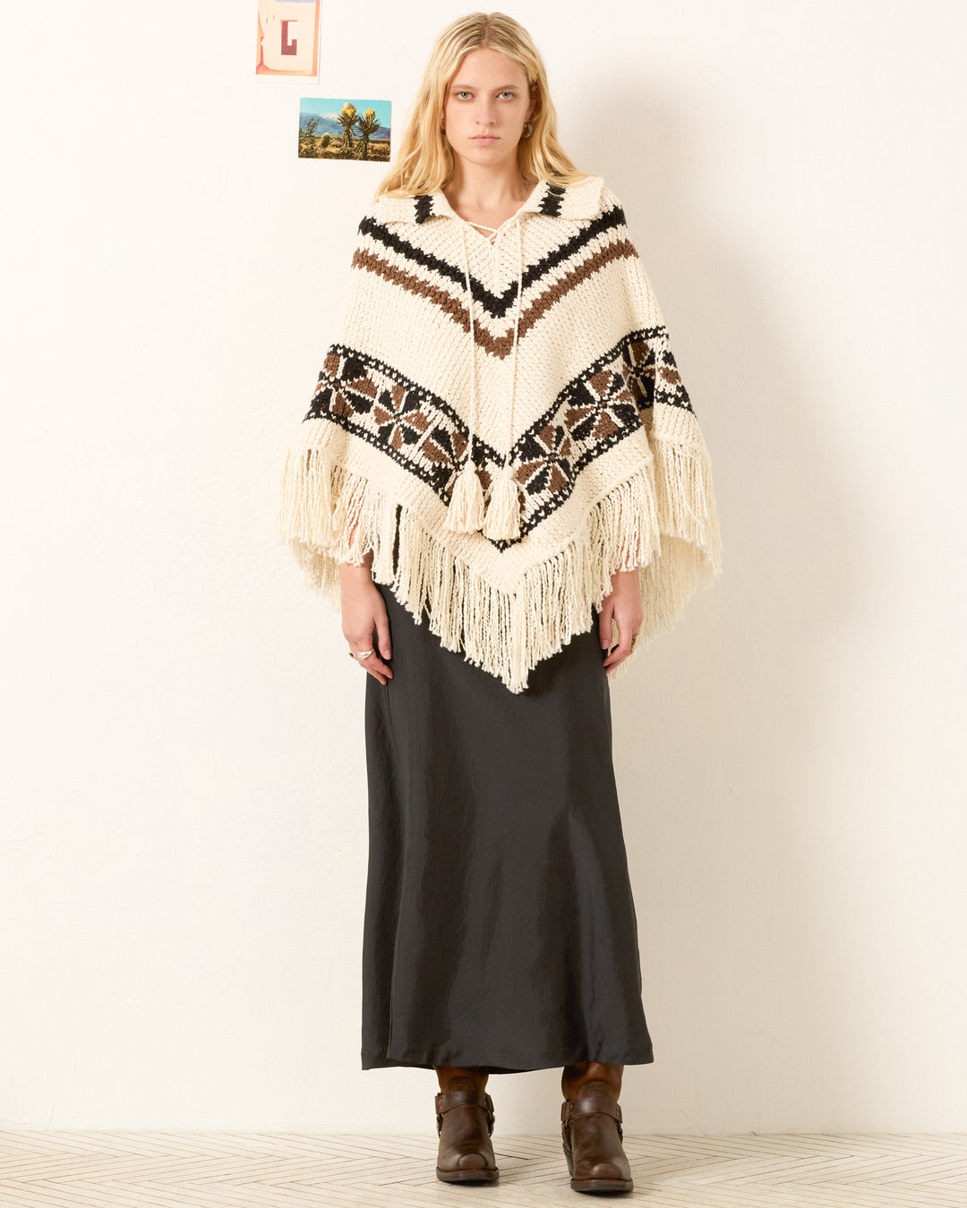 A person stands indoors wearing a black maxi skirt, brown boots, and the Faune Rawhide Knit Poncho—an artisan cream and brown poncho featuring geometric patterns and fringe.