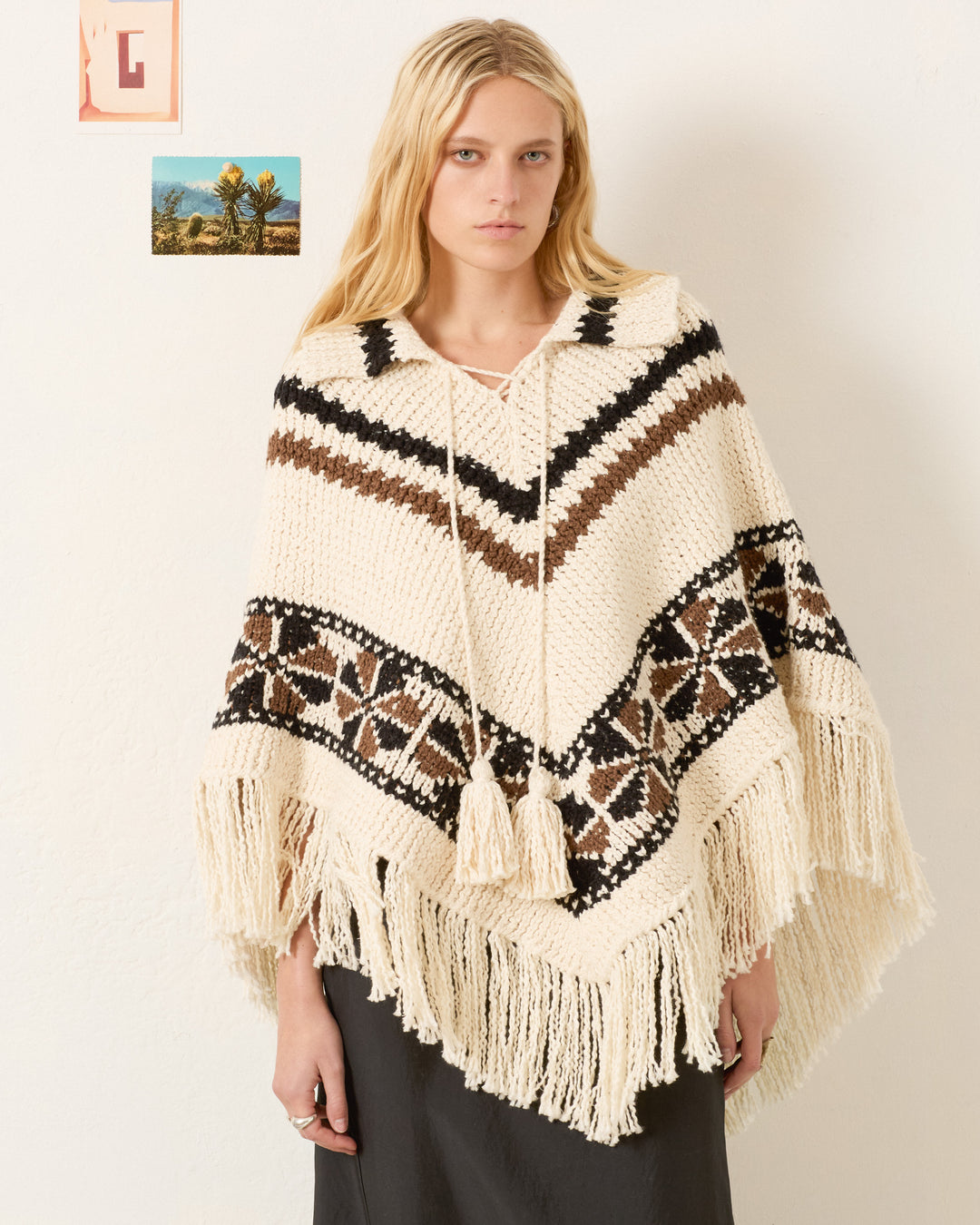 A woman with long blonde hair models the Faune Rawhide Knit Poncho, a cream-colored artisan piece with black and brown geometric patterns and fringe, as she stands before a white wall decorated with small pictures.