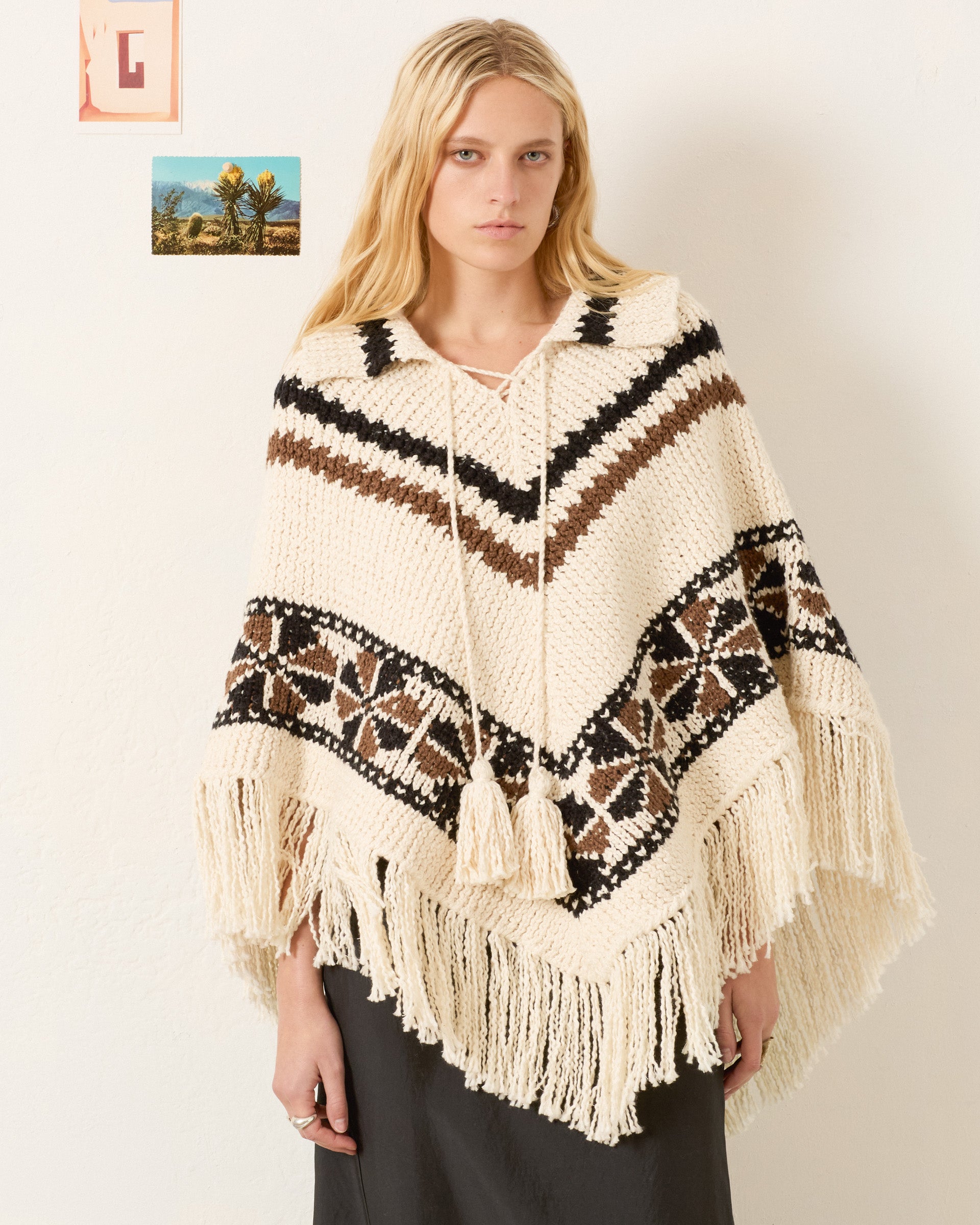 A woman with long blonde hair models the Faune Rawhide Knit Poncho, a cream-colored artisan piece with black and brown geometric patterns and fringe, as she stands before a white wall decorated with small pictures.