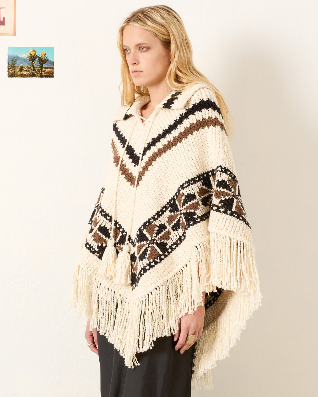 A woman with long blonde hair wears the Faune Rawhide Knit Poncho—cream-colored with brown and black geometric patterns—standing against a plain wall decorated with small pictures.