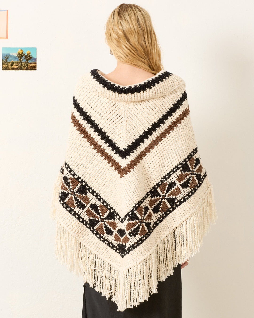 A person with long blonde hair stands with their back to the camera, wearing the Faune Rawhide Knit Poncho—a cream knit shawl featuring brown and black geometric patterns and fringe.