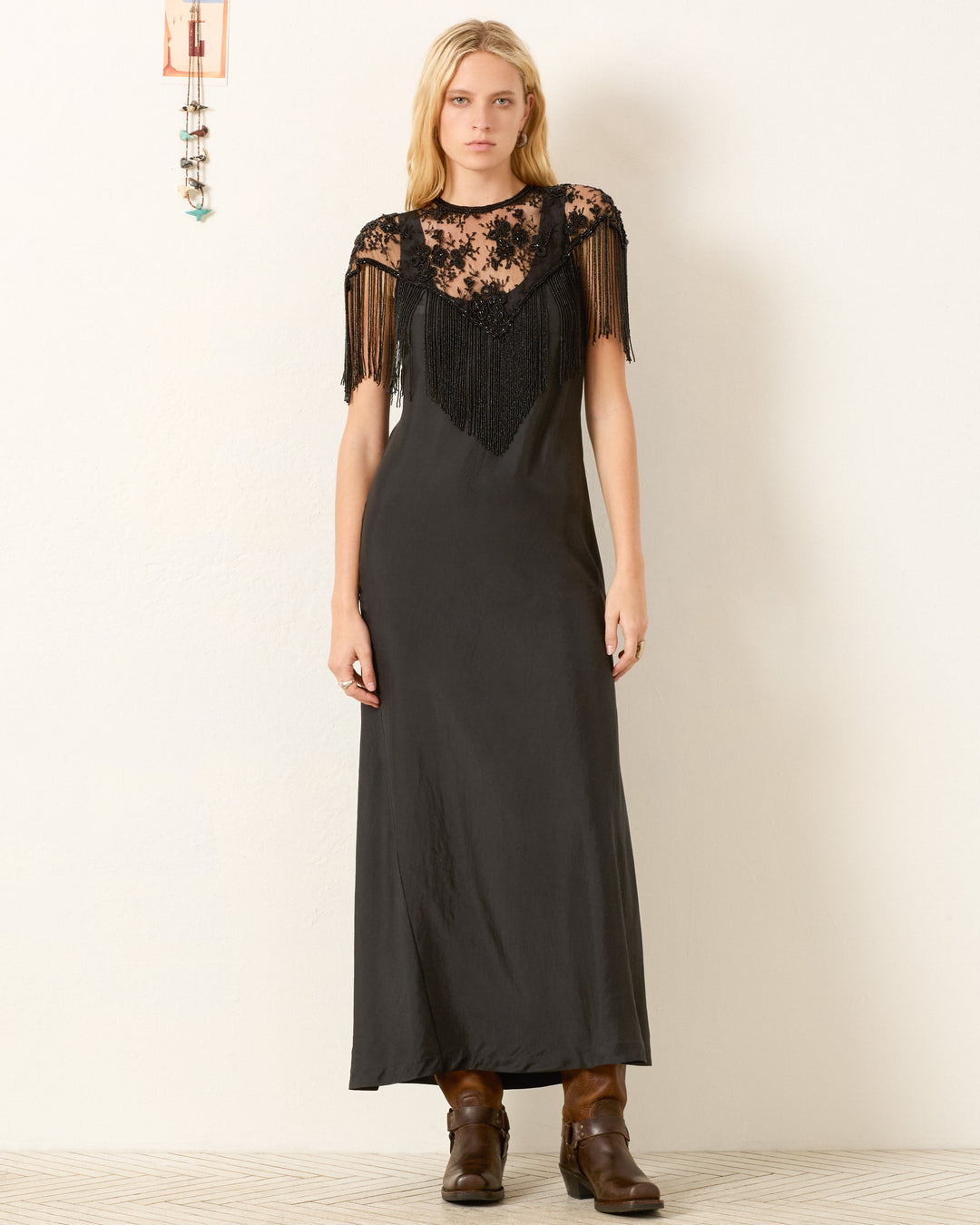 A woman stands against a plain wall wearing a long black dress with lace and fringe detailing, brown boots, minimal accessories, and the vintage-inspired Flou Night Ride Lace Capelet.