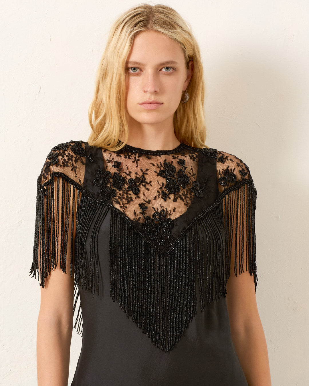 A woman with straight blonde hair wears the Flou Night Ride Lace Capelet, a vintage-inspired black piece featuring hand-beaded lace, floral embroidery, and fringe shoulders, standing against a plain light wall.