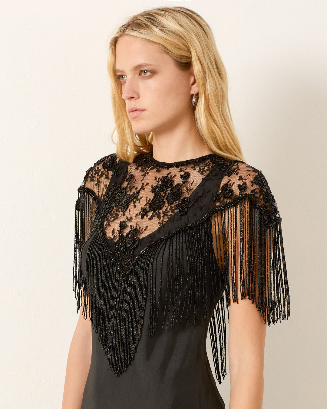 A woman with long blonde hair wears the Flou Night Ride Lace Capelet, a vintage-inspired black dress with a sheer lace neckline, fringe on the shoulders and chest, and a black lace capelet.