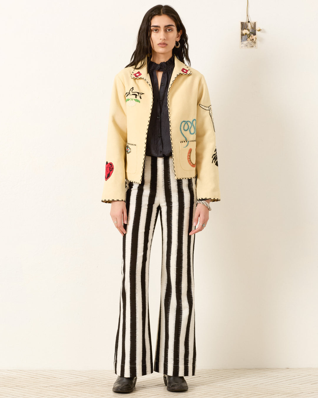 A person stands against a white background wearing the cream, beaded and embroidered Francis Rye Tattoo Jacket, a dark blouse, black and white vertical striped pants, and black shoes.