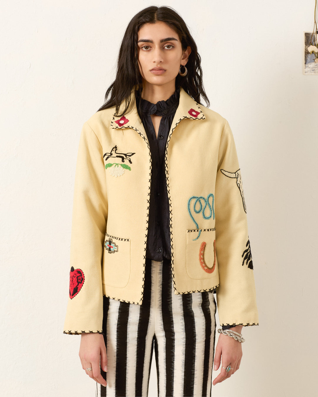 A person with long dark hair stands against a plain light background, wearing the Francis Rye Tattoo Jacket, a Western-inspired piece with colorful embroidery, paired with black-and-white striped pants.