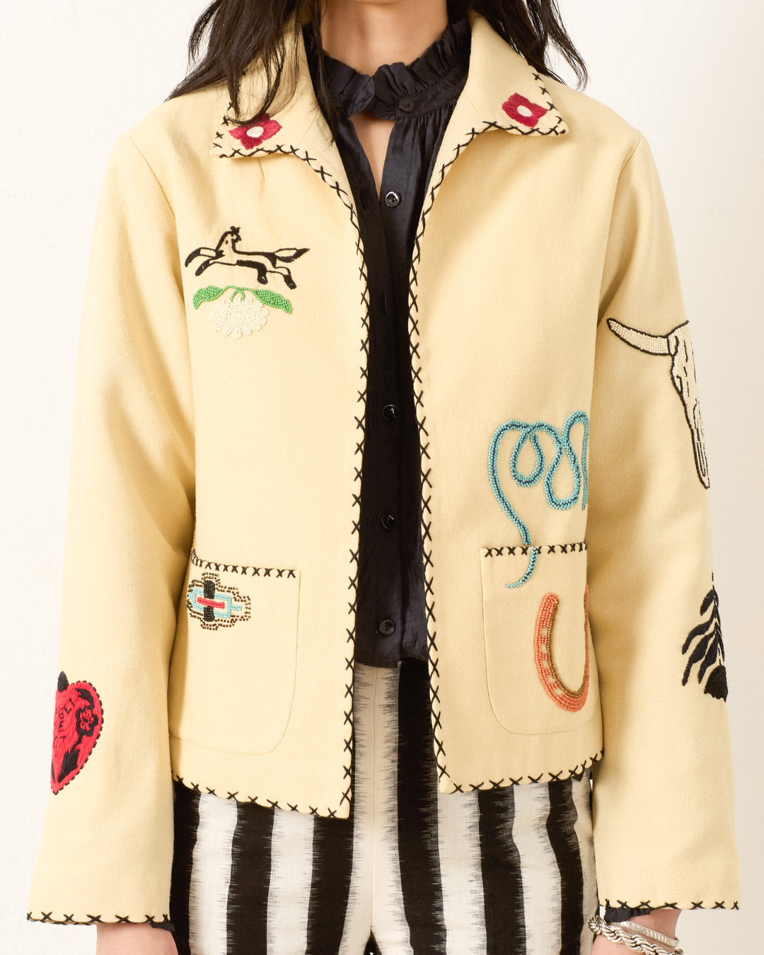 Someone wearing the Francis Rye Tattoo Jacket, a Western-inspired piece with black trim and beaded, embroidered horse, lasso, horseshoe, heart, and steer skull motifs, styled over a black shirt and striped pants.