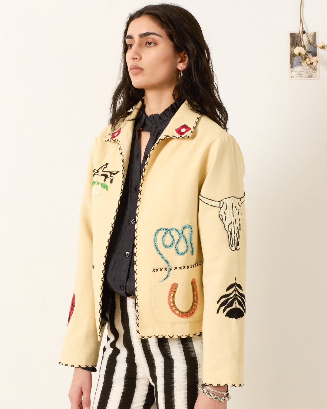 A woman wears the Francis Rye Tattoo Jacket, a cream Western-inspired piece with colorful beaded and embroidered designs, paired with black-and-white striped pants against a plain background.