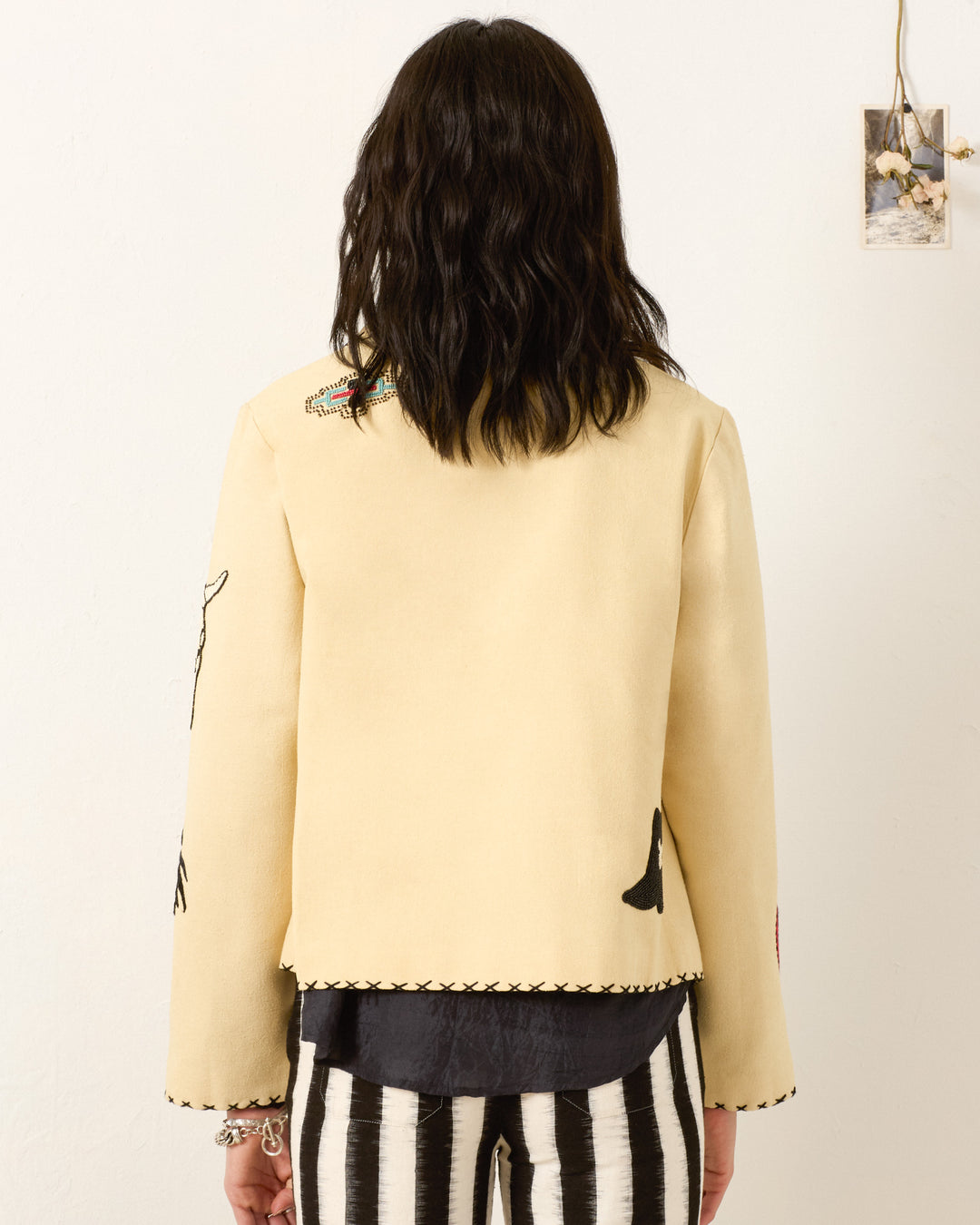 A person stands with their back to the camera in a Francis Rye Tattoo Jacket featuring stitched details and black-and-white striped pants. A small photo is pinned to the wall on the right.