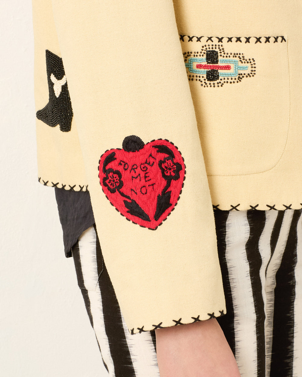 Close-up of the Francis Rye Tattoo Jacket sleeve with a red "FORGET ME NOT" heart patch and decorative stitching, worn over a black shirt and striped black-and-white pants.