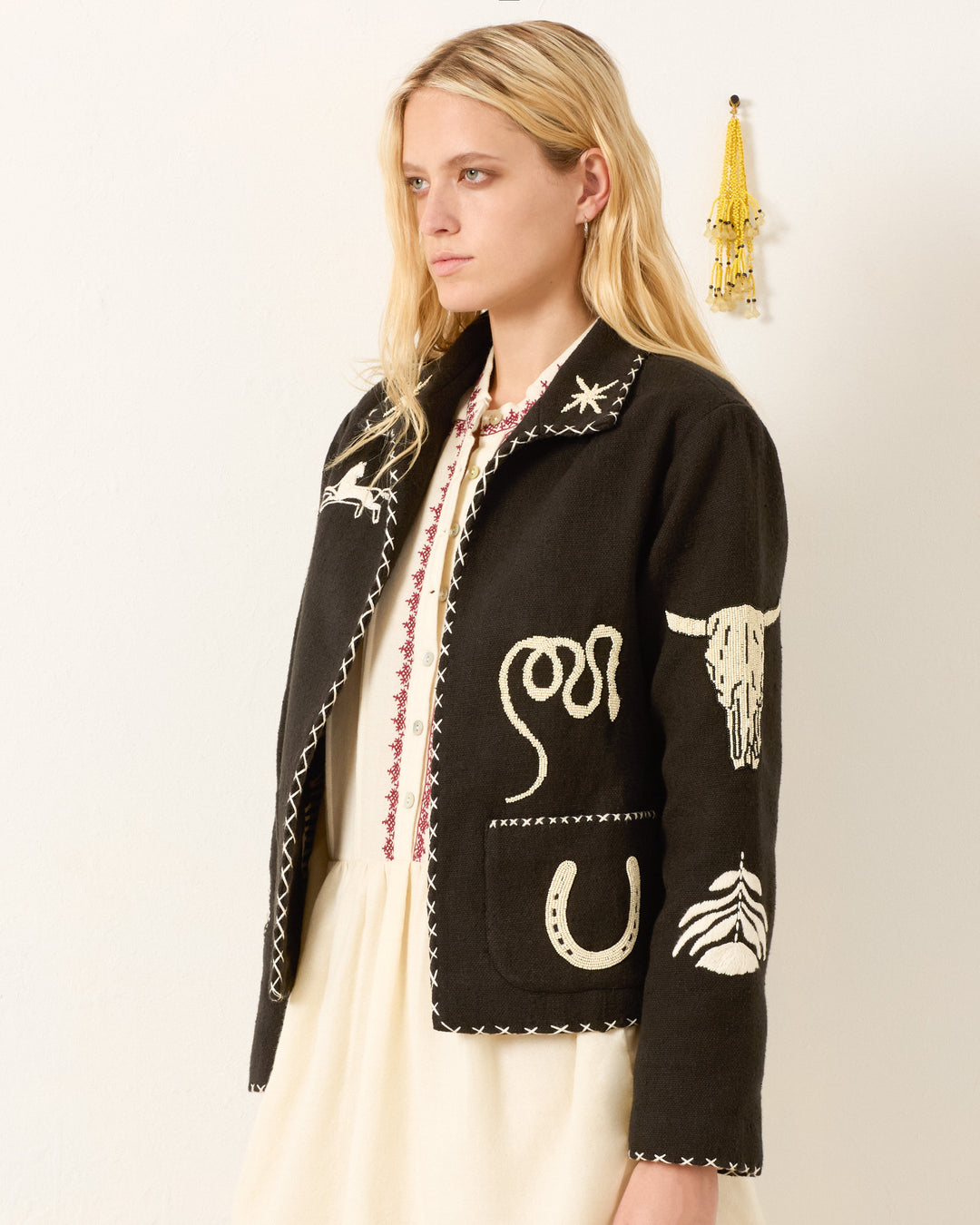 A woman with long blonde hair models the Francis Noir Tattoo Jacket, a hand-embroidered black piece with white Western-themed details, standing against a plain wall accented by a small yellow decoration behind her.