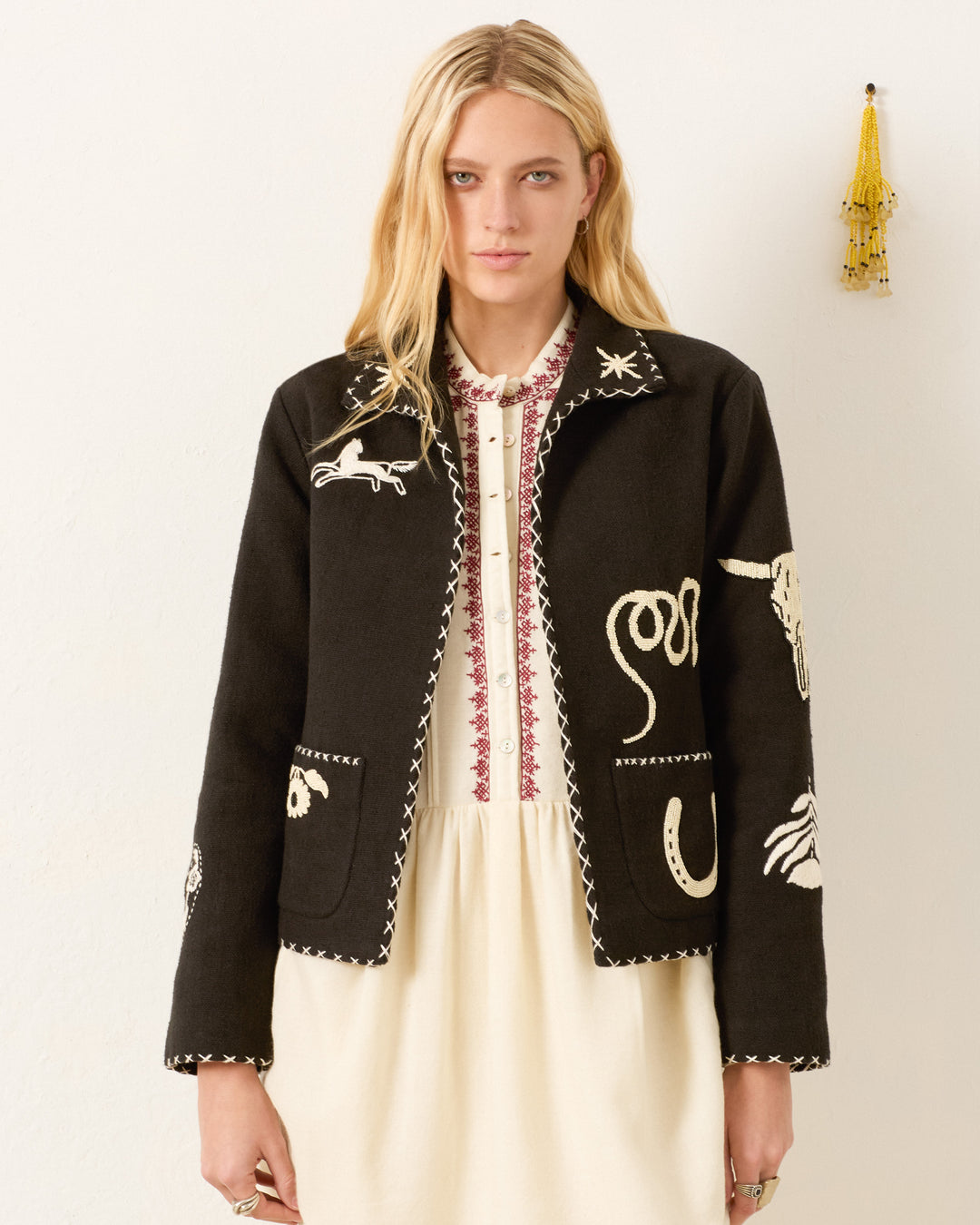 A woman with long blonde hair wears a cream dress and the Francis Noir Tattoo Jacket, a black hand-embroidered piece with white animal and abstract designs, standing against a plain wall decorated with a yellow tassel.