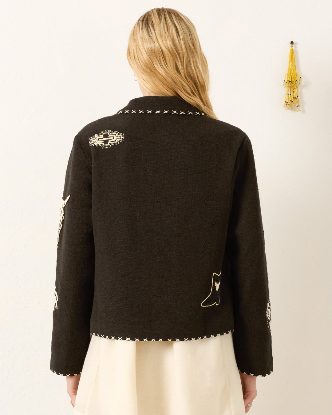 A person with long blonde hair, seen from behind against a light wall, wears the Francis Noir Tattoo Jacket—black with white hand-embroidered stitching and patches.