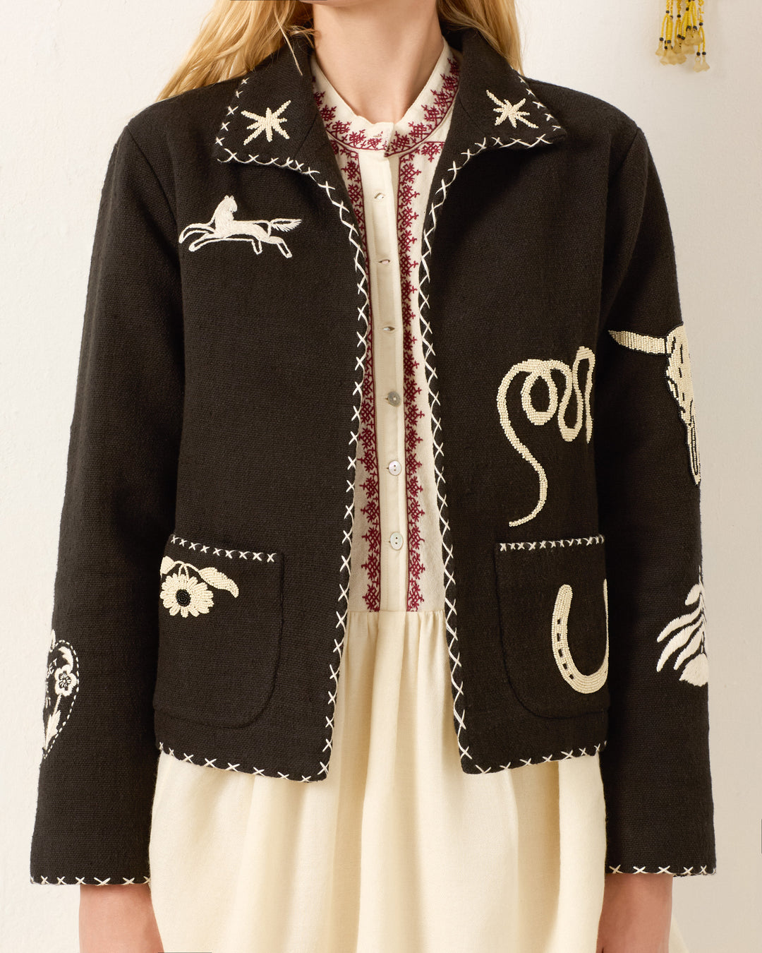 A woman wears the Francis Noir Tattoo Jacket, a black hand-embroidered piece with white Western motifs, over a cream dress with a red embroidered collar.