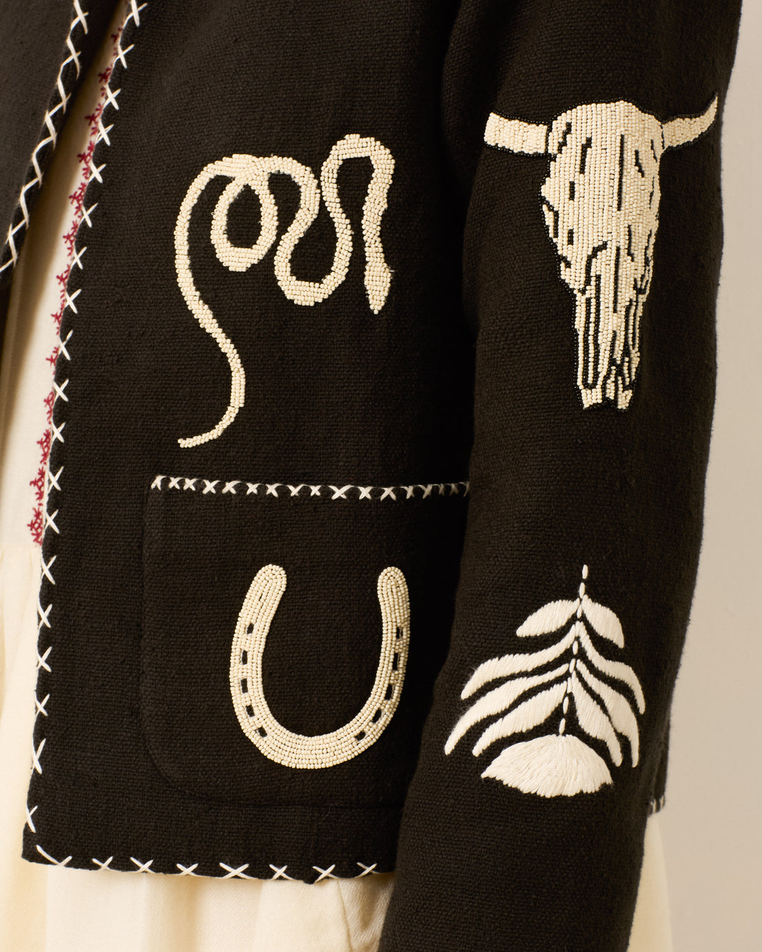 The Francis Noir Tattoo Jacket is a black hand-embroidered jacket featuring white designs—a cow skull, snake, horseshoe, feather—and white stitched edges.