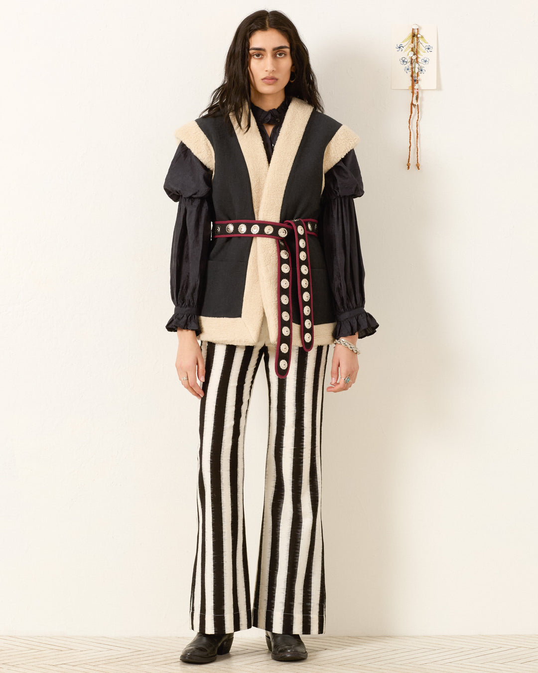 A person stands against a white wall wearing the Harrow Cactus Spur Vest, a black blouse, striped black and white pants, a maroon belt with metal accents, and black shoes.