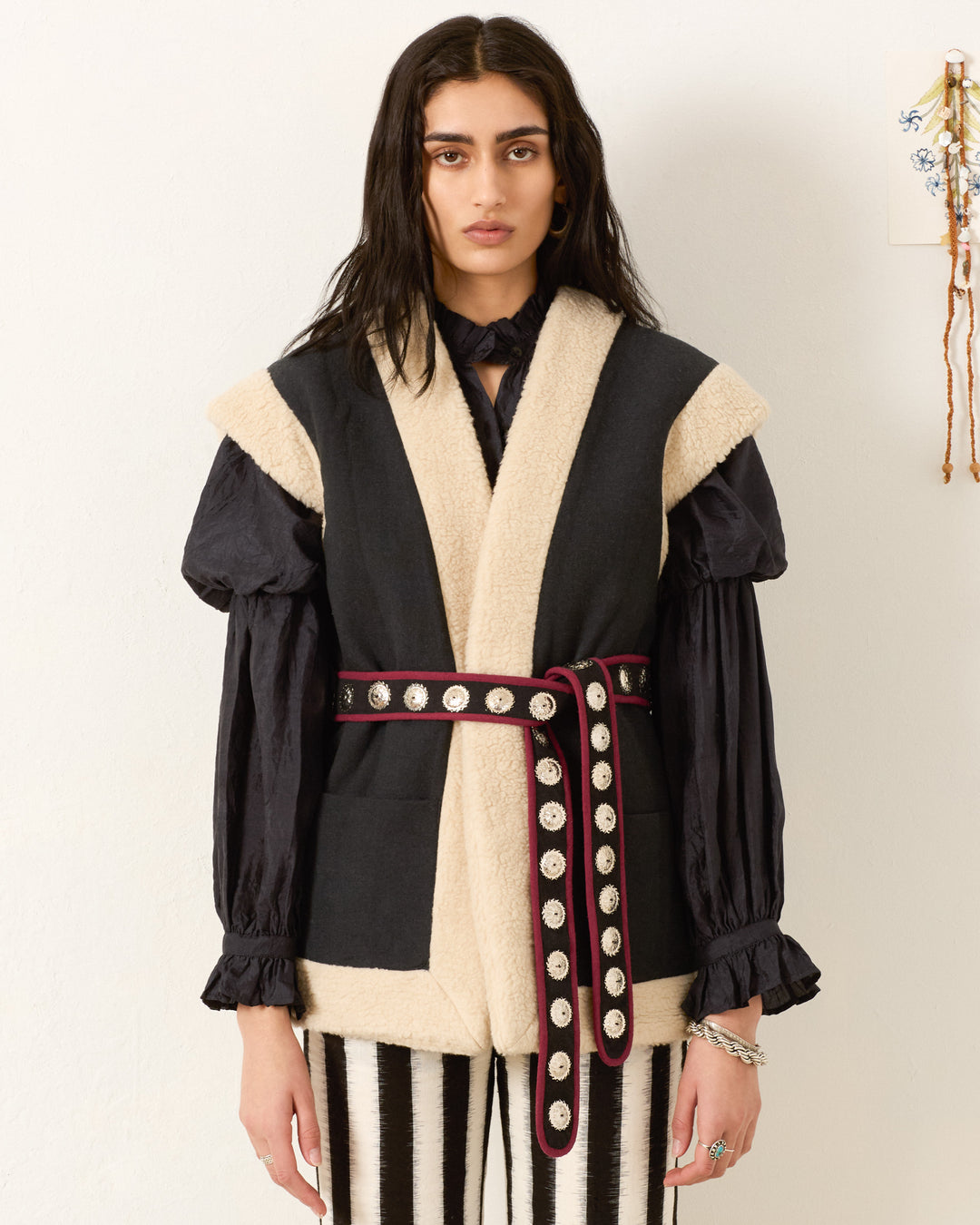 A person with long dark hair wears a black blouse, the Harrow Cactus Spur Vest with a wide belt featuring circular metal accents, and black and white striped pants.