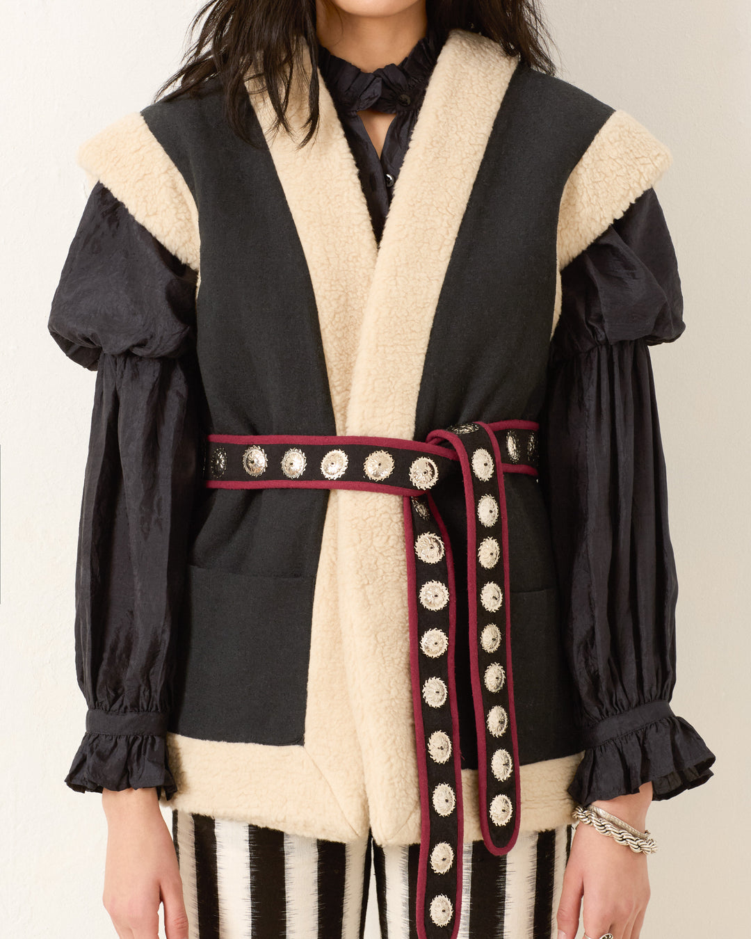 A person wears a black blouse with puffed sleeves, the Harrow Cactus Spur Vest with textured trim, a decorative belt, and striped black and white pants.