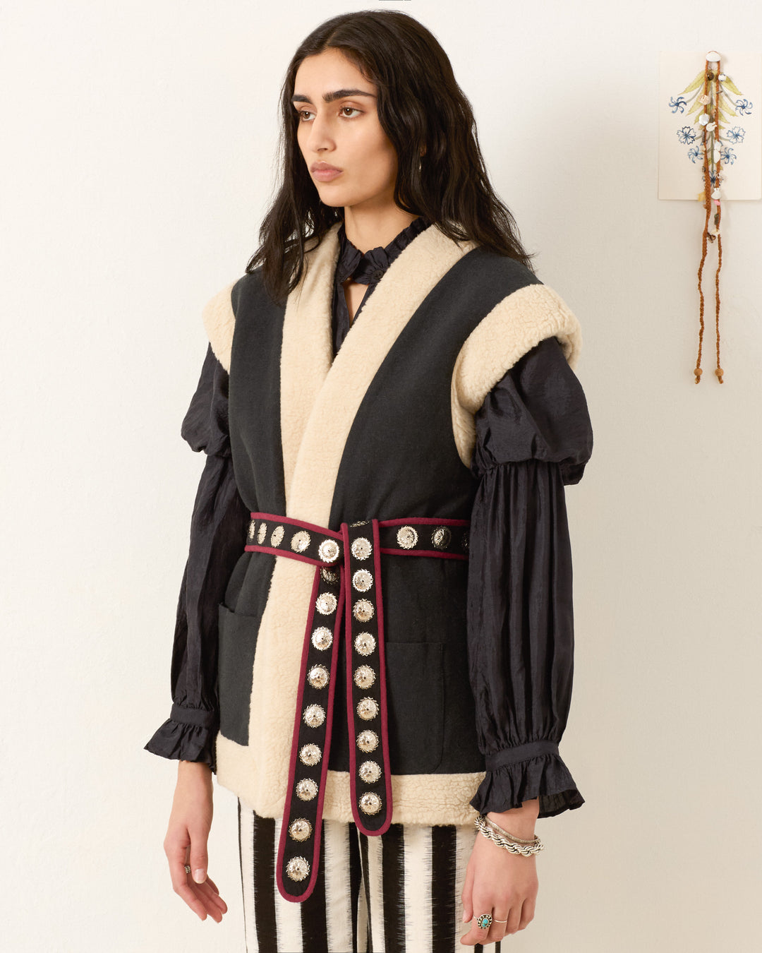 A person wearing a black puff-sleeve blouse, the Harrow Cactus Spur Vest in cream and black with silver embellishments, and vertically striped pants stands against a plain wall with a small decoration.