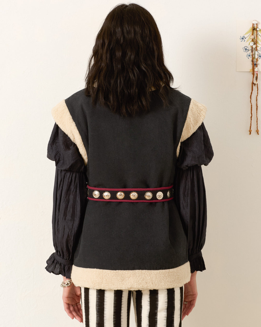 A person stands facing away, wearing the Harrow Cactus Spur Vest with beige trim, a black puff-sleeve shirt, decorative belt, and black-and-white striped pants against a plain wall.