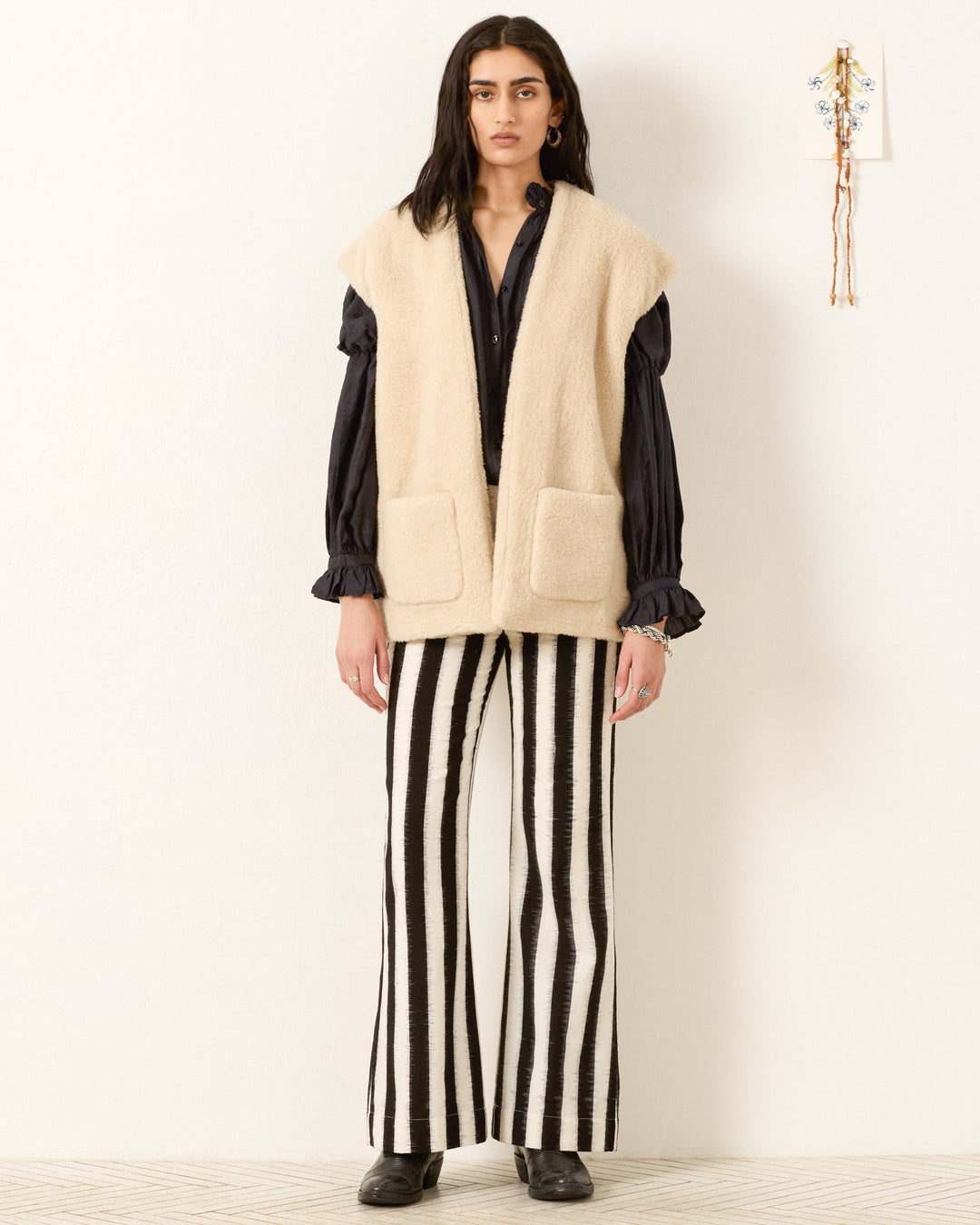 A person stands against a white wall wearing the beige Harrow Cactus Spur Vest, a black blouse, black-and-white striped pants, and black shoes.