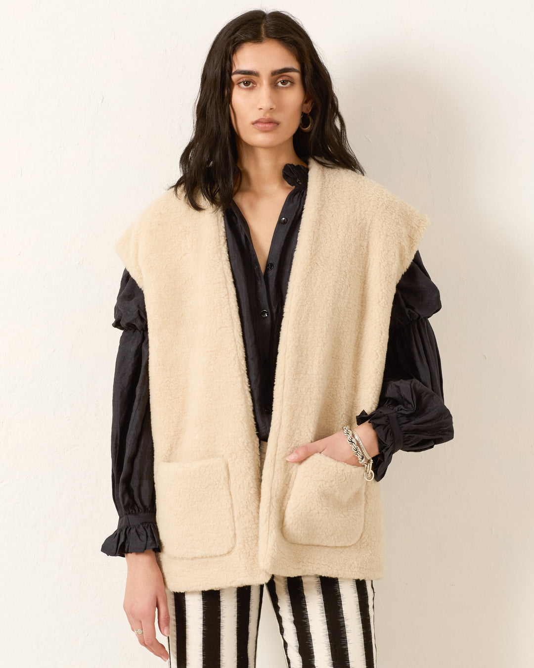 A woman stands against a plain wall wearing the Harrow Cactus Spur Vest over a dark blouse and black-and-white striped pants.