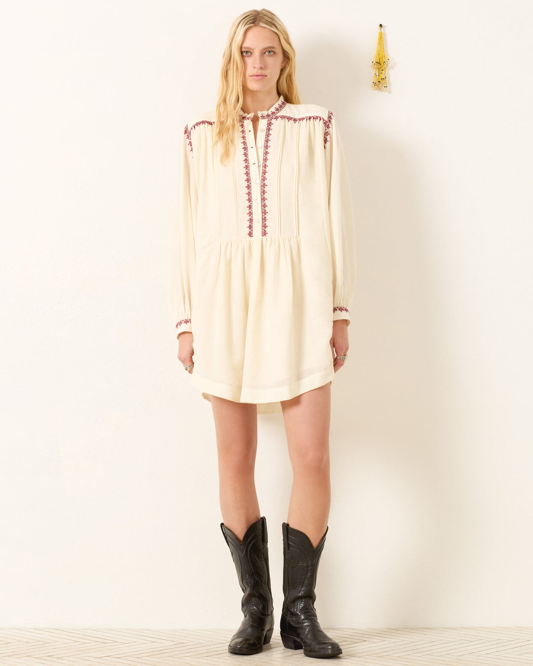 A woman stands against a white wall wearing the Jolie Snow Beam Dress—a cream, hand-embroidered long-sleeve dress with red detailing—and black cowboy boots. A small hanging decoration is beside her on the wall.