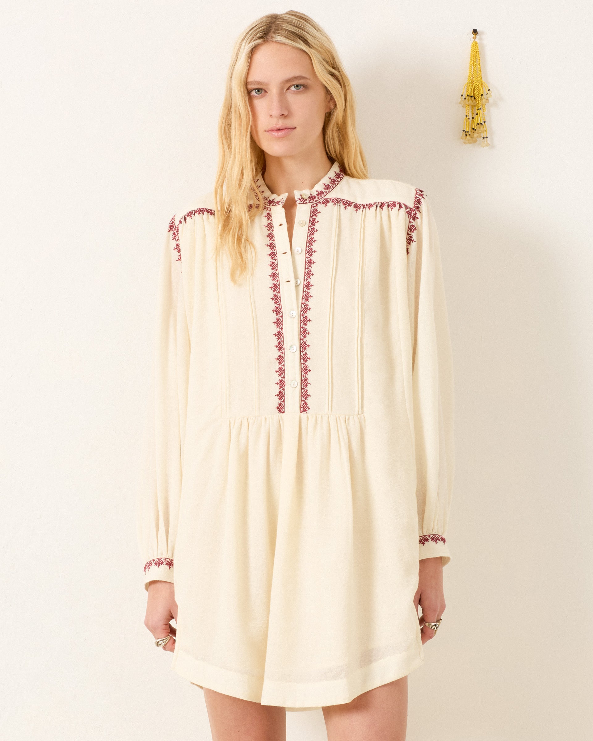 A woman stands by a plain wall, wearing the Jolie Snow Beam Dress, a long-sleeve wool shirt dress featuring hand-embroidered red details on the band collar, shoulders, and cuffs. A yellow tassel decorates the wall nearby.