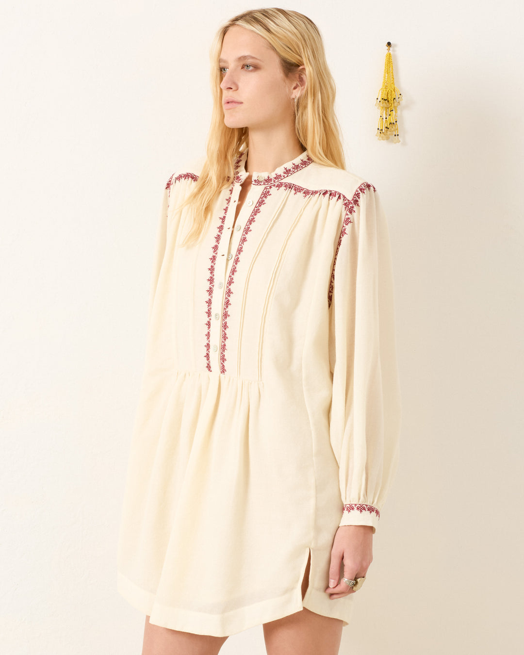 A woman stands against a white wall wearing the Jolie Snow Beam Dress, a cream hand-embroidered wool shirt dress with red accents and a band collar. Yellow tassel decor hangs on the wall beside her.