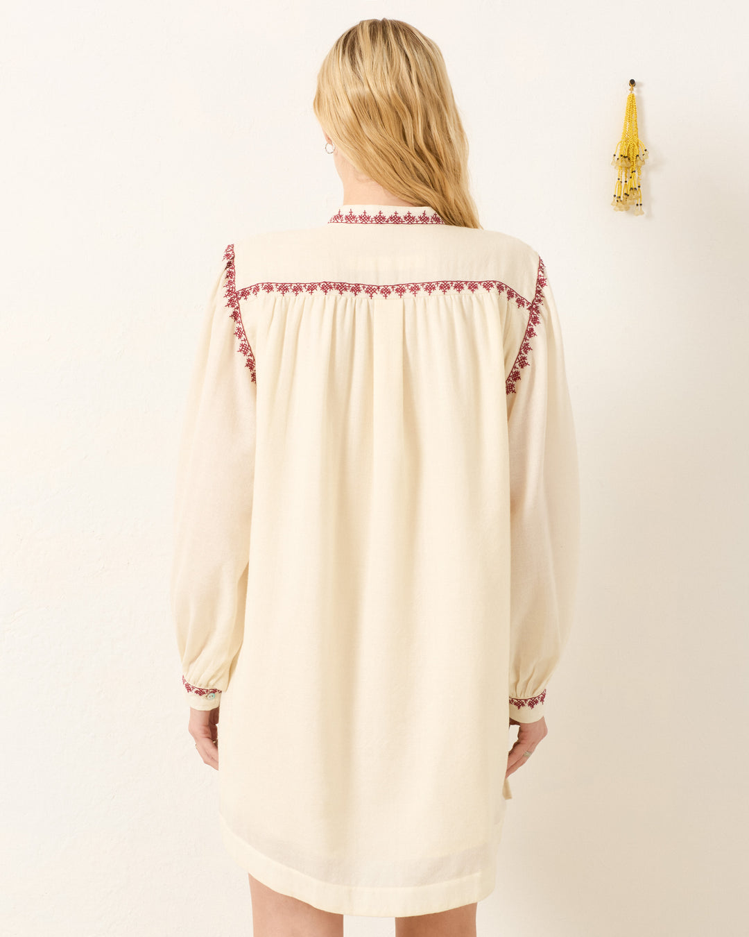 A woman with long blonde hair faces away, wearing the Jolie Snow Beam Dress—a cream wool shirt dress with a band collar and red hand-embroidered yoke and sleeves—standing near a white wall with a small yellow wall hanging.