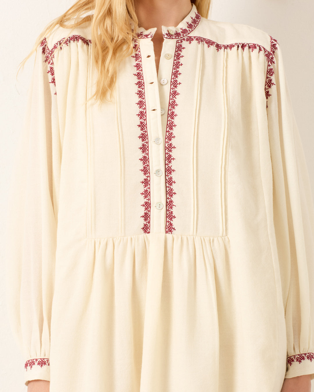 A woman wearing the Jolie Snow Beam Dress, a cream-colored long-sleeve blouse with red hand embroidery, front buttons, and a chic band collar.