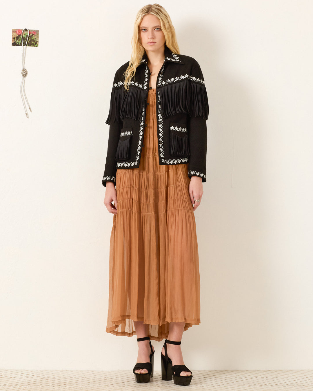 A woman stands against a plain wall wearing the Jude Raven Fringe Jacket, a black wool piece with artisan hand embroidery, styled over a long light brown dress and black platform heels.