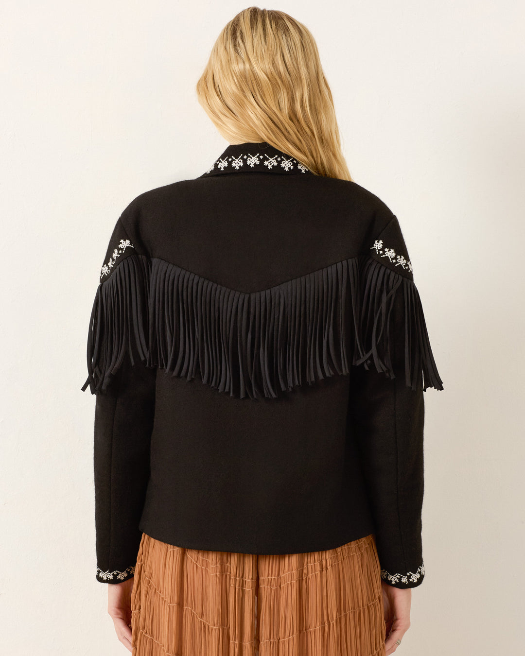 Someone wearing the Jude Raven Fringe Jacket, featuring wool fabric and hand-embroidered details on the collar and sleeves, styled with a brown pleated skirt.