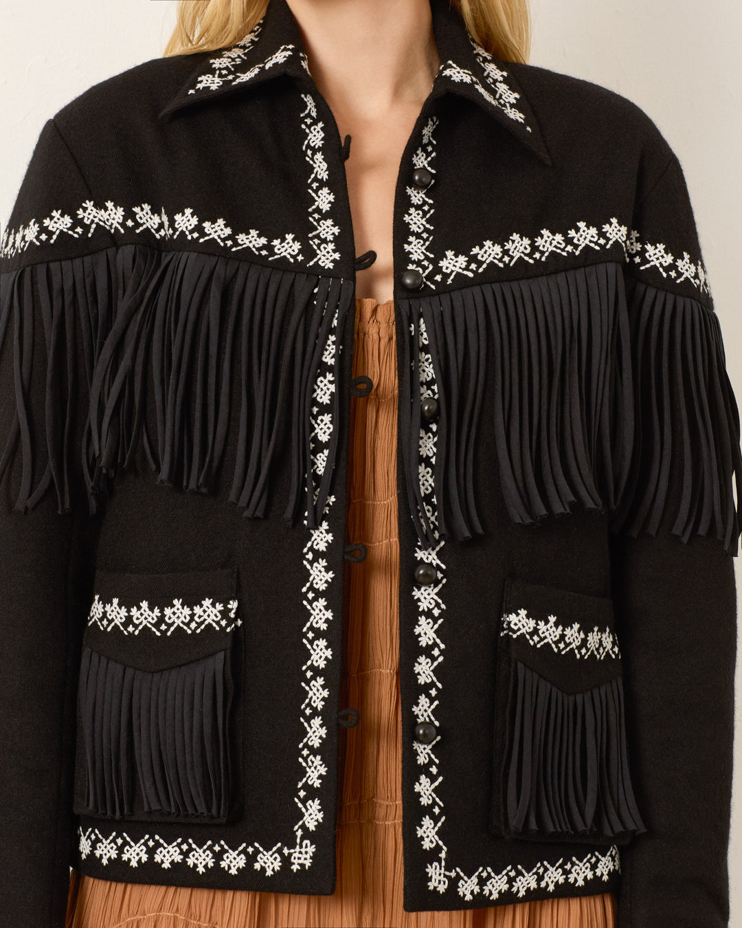 A person wearing the Jude Raven Fringe Jacket, a stylish wool jacket with decorative white hand embroidery and fringe, over a brown top.