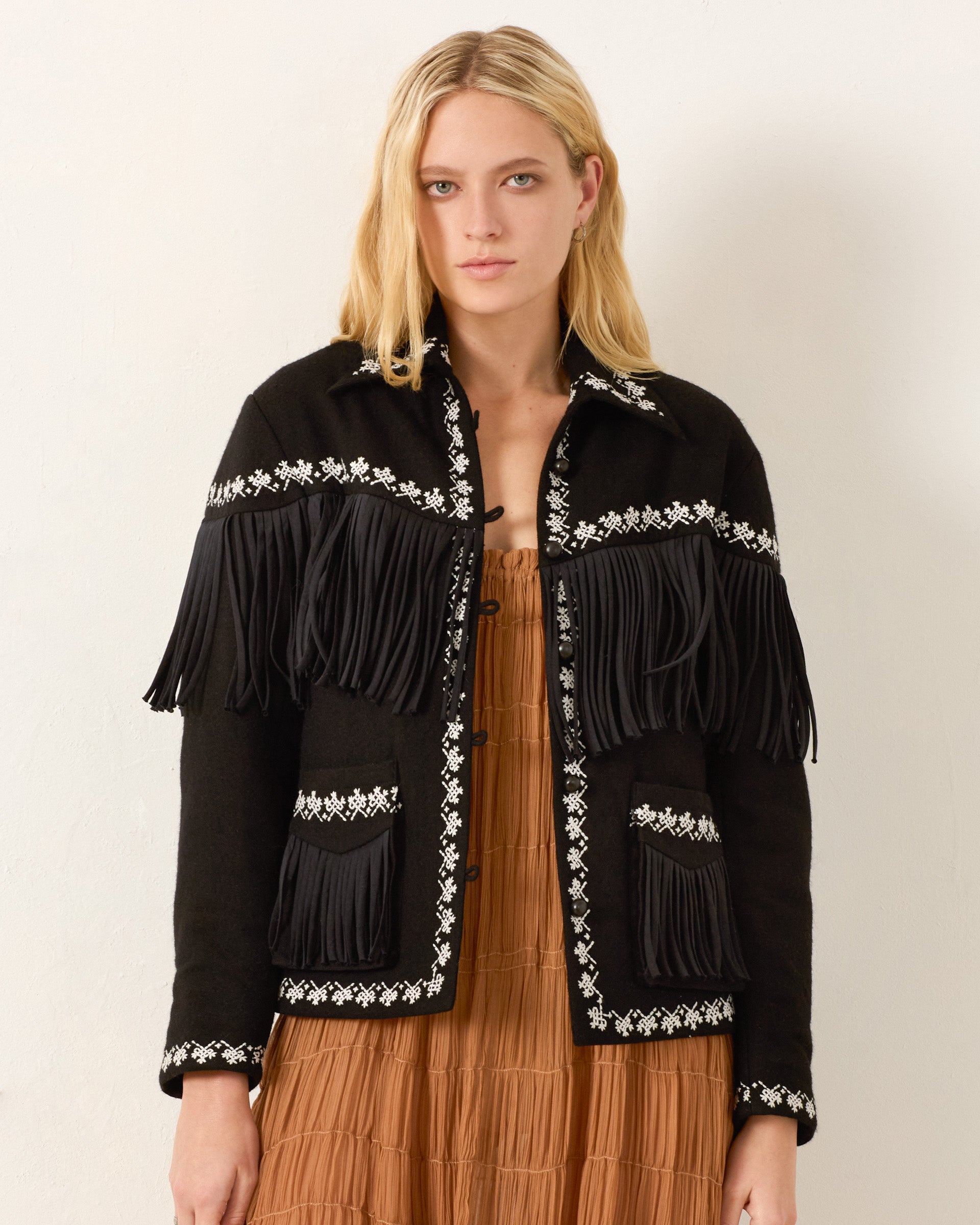 A woman wears the Jude Raven Fringe Jacket, an artisan wool piece with hand embroidery, over a brown dress against a plain light background.