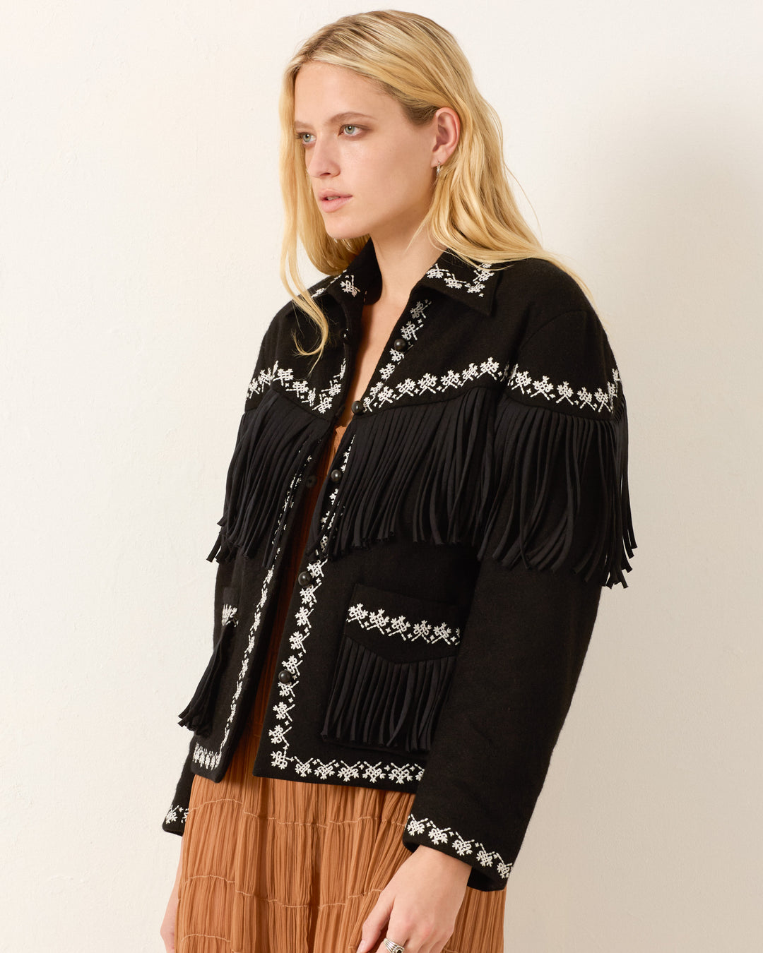 A woman stands against a plain light background, wearing the Jude Raven Fringe Jacket—black wool with white floral hand embroidery and fringed edges—layered over a brown dress.