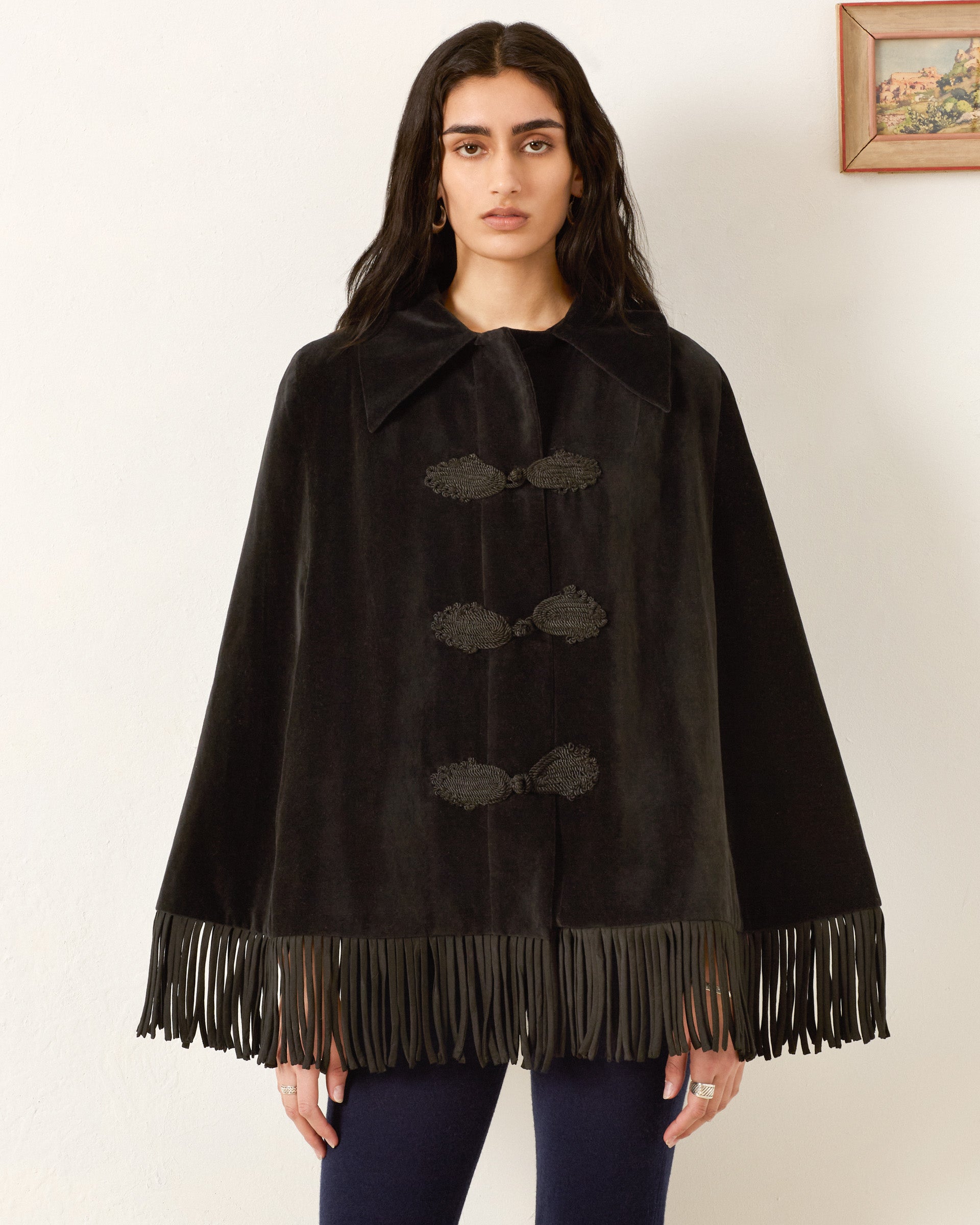 A person stands against a white wall wearing the Kensington Onyx Fringe Cape with decorative clasps and dark pants. A small framed artwork hangs on the wall.